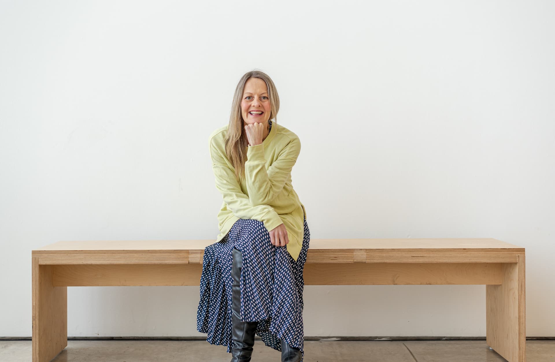Clare Inkster sitting on a plywood bench, smiling, in a pale yellow sweatshirt