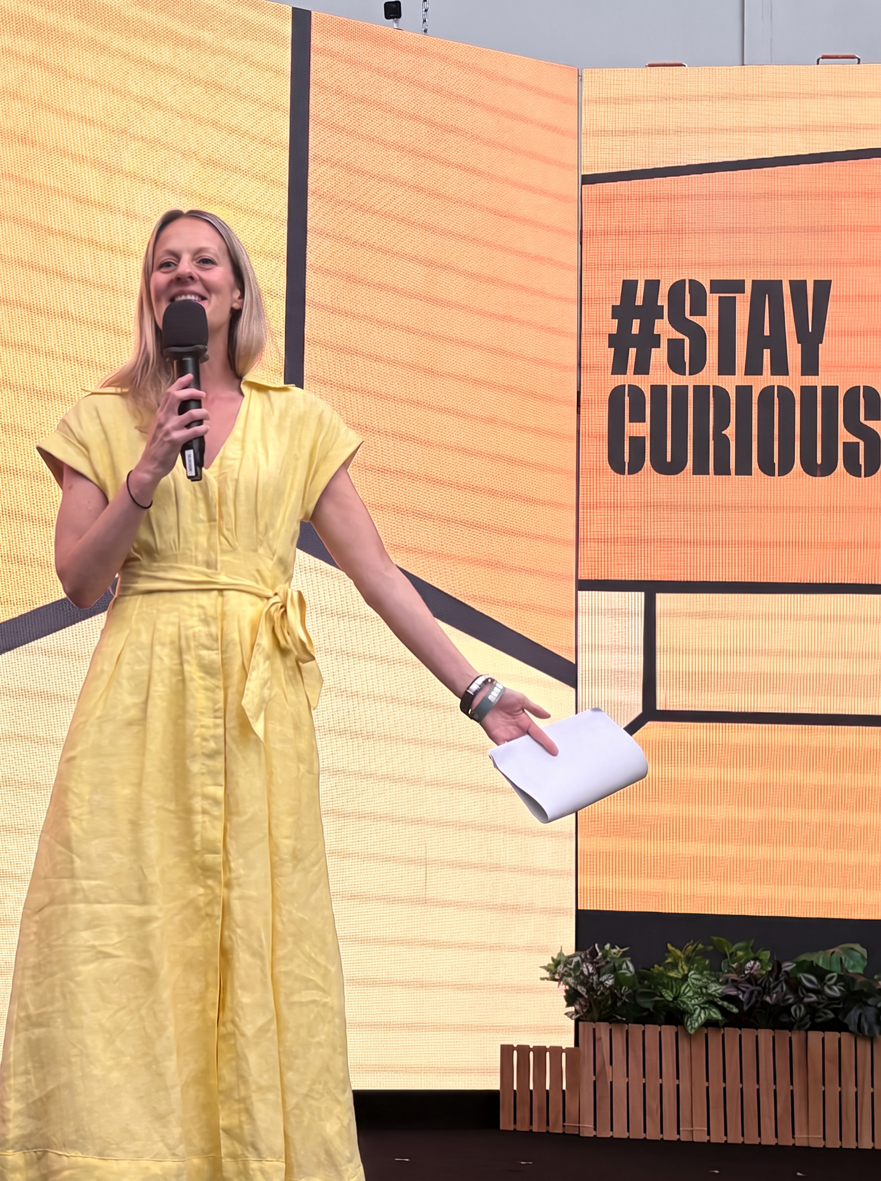 Clare Inkster speaking on stage with a microphone in front of a large orange screen reading #staycurious