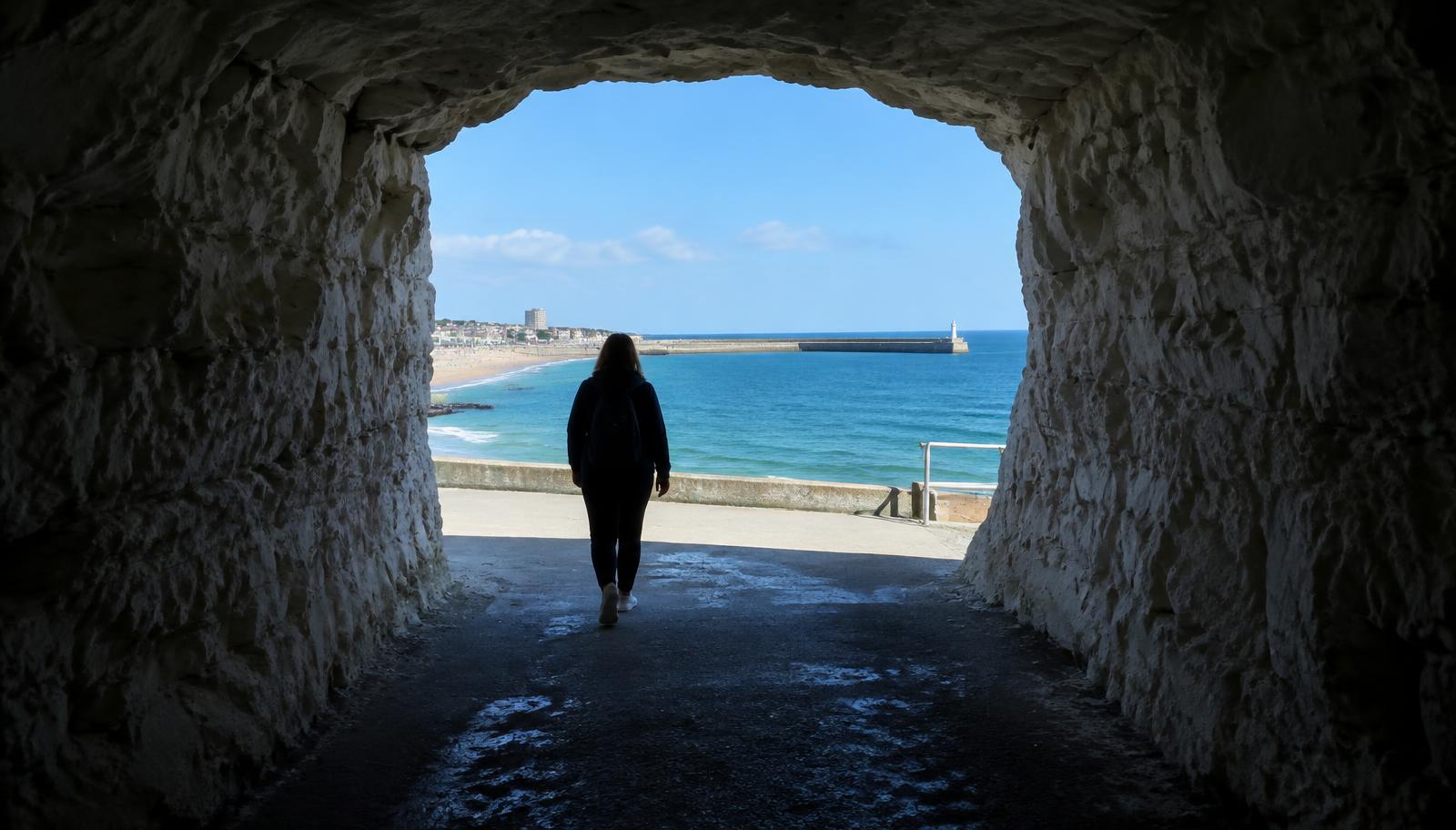 Looking out of a dark chalk tunnel onto a bright blue sea, with someone walking towards the light